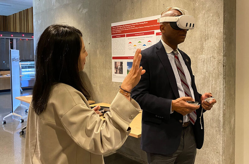Two people interacting with virtual reality equipment in a modern room; one wearing a VR headset and holding controllers, while the other gestures with one hand. A red and white poster with diagrams is displayed on the concrete wall behind them.