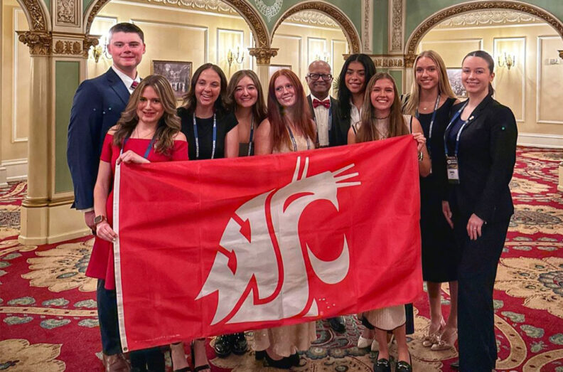 A group of conference attendees stand together holding a large crimson Washington State University flag with the cougar logo in an ornate room featuring patterned carpet, decorative arches, and gold accents during the Washington Tourism Conference.