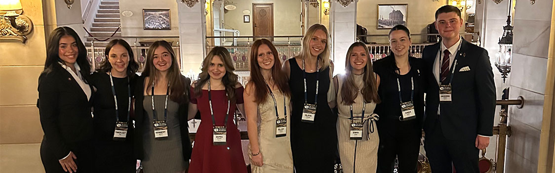 A group of hospitality business management students dressed in professional attire stand together in an ornate lobby with chandeliers and decorative railings during the 2025 State of Washington Tourism Conference.