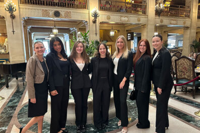 A group of students wearing conference badges pose together in a grand interior space with ornate railings, chandeliers, and framed artwork during the State of Washington Tourism Conference.