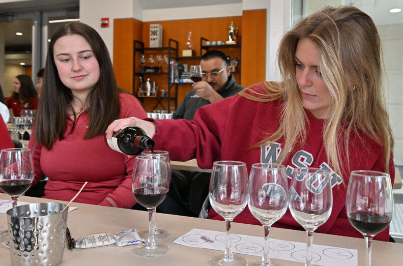 Students wearing a WSU sweatshirts pour red wine into tasting glasses during a wine tasting session, with multiple glasses and a tasting sheet on the table.