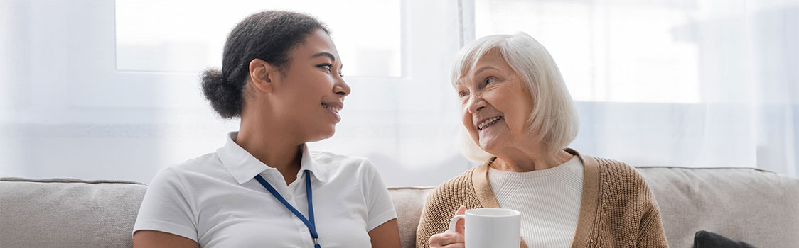 A healthcare professional wearing a white polo shirt with a blue lanyard sits on a couch next to an older adult in a tan sweater holding a white mug, with bright windows in the background.