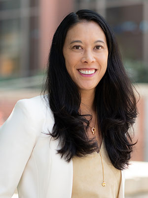 Hana Johnson wearing a cream blouse and white blazer, with long dark wavy hair, standing outdoors in front of softly blurred campus buildings.