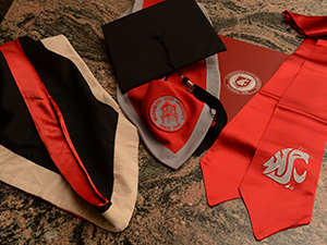 WSU graduation items arranged on a table, including a cap, gown hood, red stole with the Cougar logo, and a folder with the WSU seal.