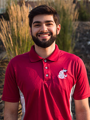 Alex Soberanes standing outdoors in sunlight, wearing a crimson WSU polo shirt with the Cougar logo, with tall grasses in the background.