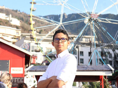 Elio Arjona standing with arms crossed in a sunny outdoor plaza, with a Ferris wheel, chalet-style buildings, and mountain slopes in the background.