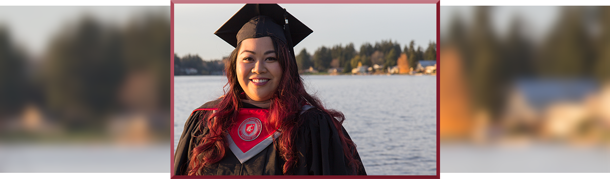 Hazelmae Overturf in a graduation cap and gown with crimson‑tinted hair, standing outdoors near a body of water at sunset, wearing a WSU graduation stole.