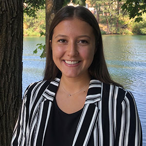 Josie Orvik standing outdoors by a lakeside, wearing a black top and striped jacket with trees and water in the background.