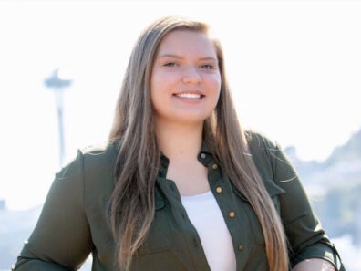 Margaret Bader standing outdoors in sunlight, wearing a dark green button‑up shirt over a light top, with an urban skyline softly visible in the background.