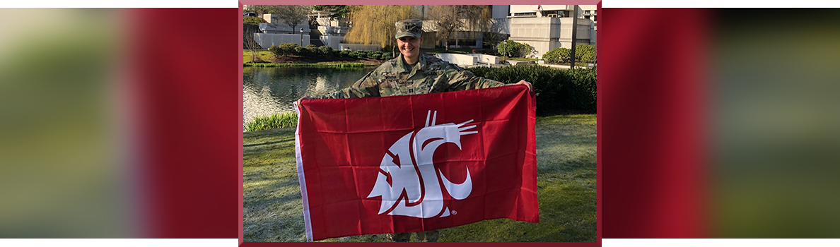 Capt. Vladi Ivanova in military uniform holding a large crimson Cougar flag outdoors beside a pond, with buildings and landscaped greenery in the background.