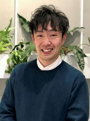 Yoshiki Shimizu standing indoors against a backdrop of leafy plants, wearing a dark sweater over a white collared shirt.