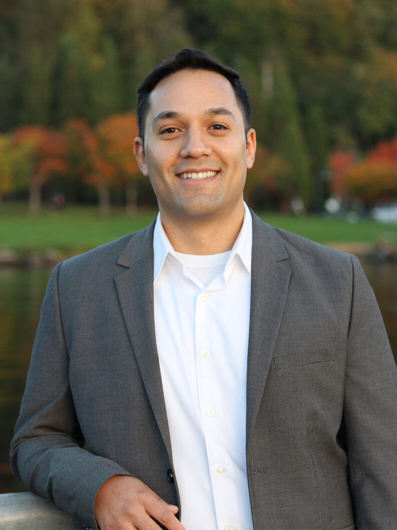 Esteban Martinez standing outdoors beside a waterfront, wearing a gray blazer over a white shirt, with autumn trees and calm water in the background.