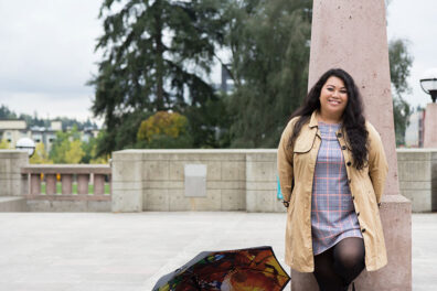 Hazelmae Overturf leaning against a stone column outdoors, wearing a light coat and plaid dress, with an open umbrella nearby and trees in the background.