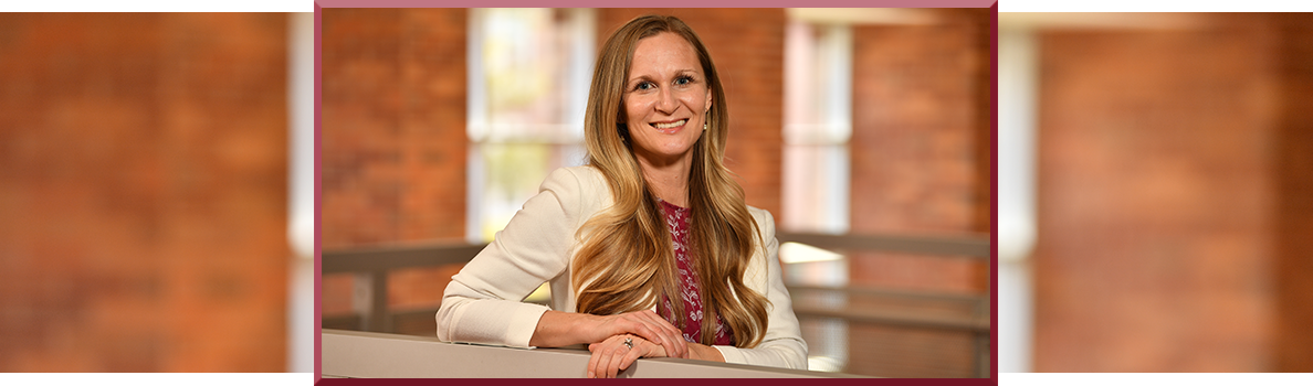 Kathleen Harris standing indoors against a warm brick background, wearing a white blazer over a patterned burgundy top with long wavy blonde hair