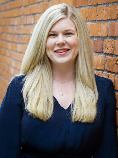 Kaycee LeCong standing beside a brick wall, wearing a navy top and a simple pendant necklace, with long blonde hair falling over her shoulders.