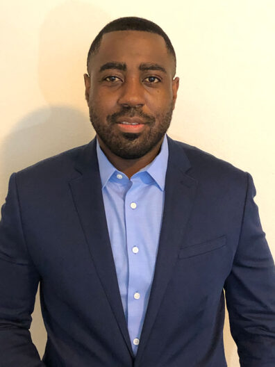 Leland Johnson wearing a navy suit jacket over a light blue button‑up shirt, standing against a plain light-colored wall.