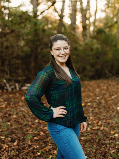 Lindsey Bennett standing in a wooded area during autumn, wearing a green plaid blouse and jeans, with one hand on her hip among fallen leaves and trees.