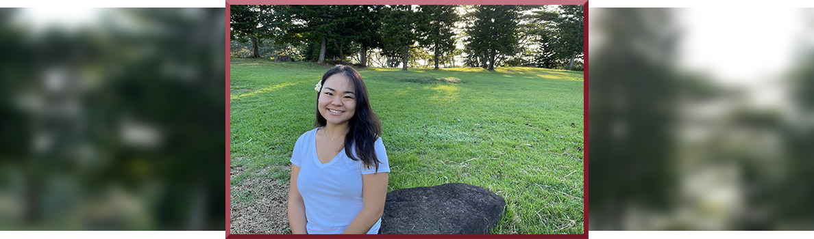 Naomi Kim sitting on a large rock in a sunny park, wearing a light blue shirt, with open grassy fields and tall trees behind her.