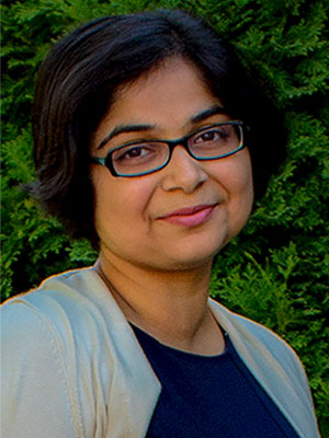 Amrita Lahiri outdoors in a light beige cardigan over a dark top, with short dark hair and green foliage behind her.