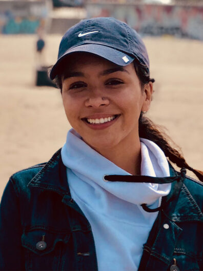 AJ Diaz outdoors on a beach, wearing a navy baseball cap, white hoodie, and denim jacket, with sunlit sand and distant structures in the background.