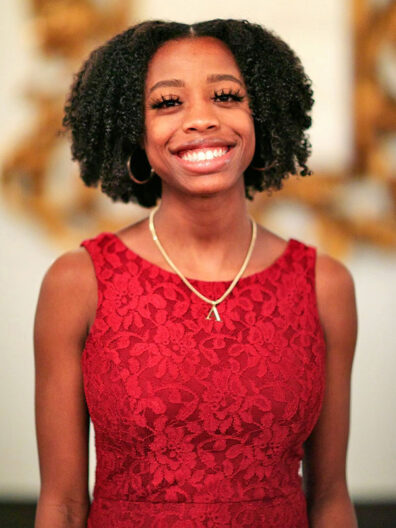 Amiyah Johnson wearing a sleeveless red lace dress and a gold necklace, standing indoors with softly blurred decorative elements in the background.