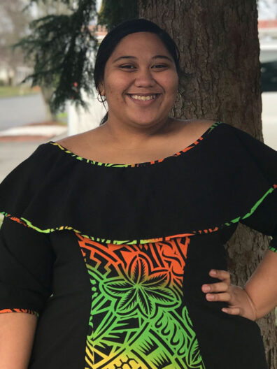 Folasa Faaitu standing outdoors beside a tree, wearing a black dress with colorful green, orange, and yellow patterned accents and an off‑shoulder neckline.