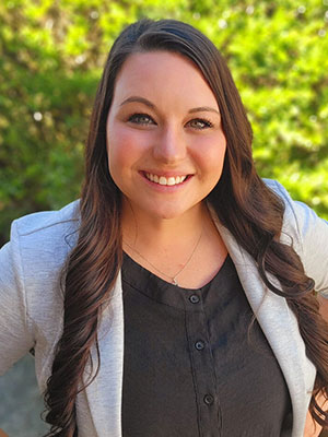 Crystal Campbell standing outdoors in sunlight, wearing a light blazer over a black top, with long wavy hair and greenery in the background.