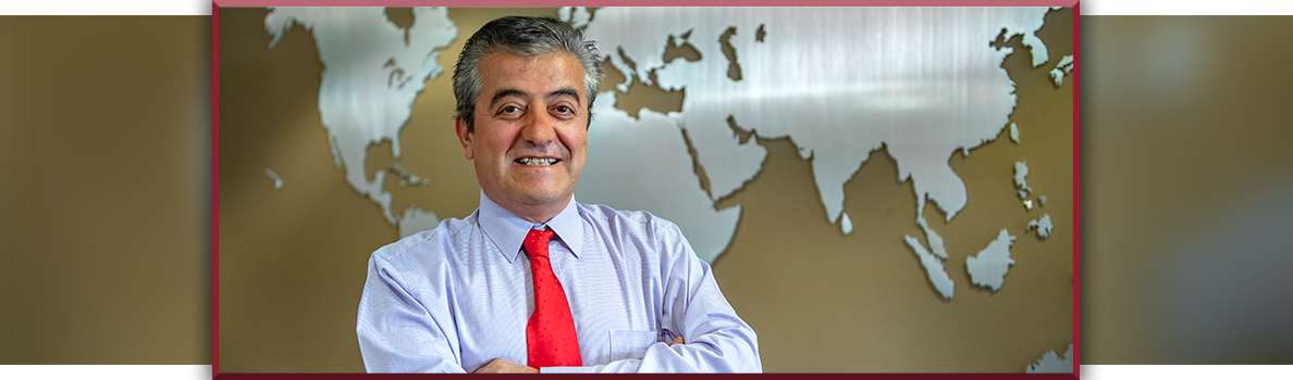 Dogan Gursoy standing with arms crossed, wearing a light purple dress shirt and bright red tie, posed in front of a world map wall display.