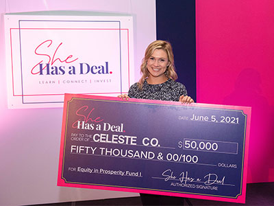 Emma Claire Spring holds an oversized $50,000 She Has a Deal award check, standing in front of a pink and white event backdrop for the hotel pitch competition.