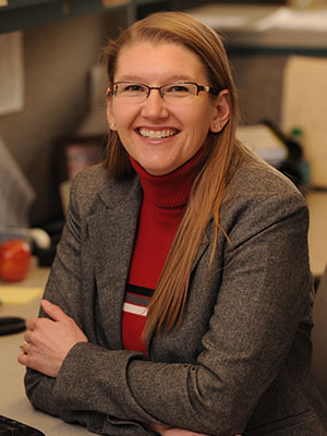 Lisa Hunter seated at an office desk, wearing a gray blazer over a red sweater, with long straight hair draped over one shoulder.