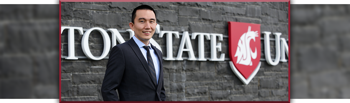 Oscar Chi standing in front of a gray stone wall with the WSU logo and lettering, wearing a dark suit with a light blue shirt and black tie.