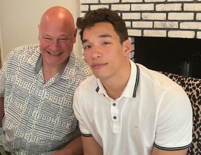 Brighton Maughan and Drew Maughan seated indoors, one wearing a patterned button‑up shirt and the other a white polo, with a brick wall and leopard‑print chair behind them.