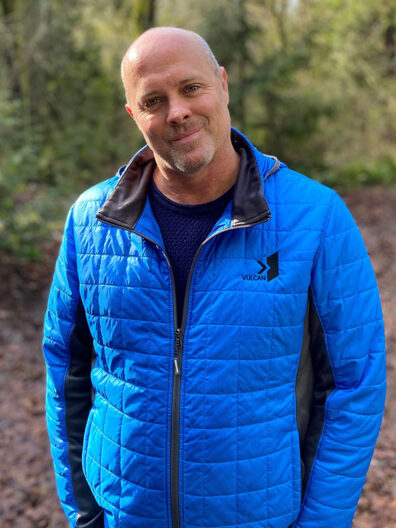 Tim Mulligan wearing a bright blue quilted Vulcan jacket and navy shirt, standing on a wooded trail with soft greenery in the background.