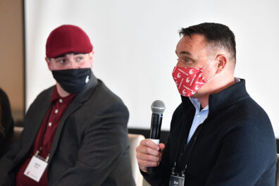 Brian Lewis speaks into a handheld microphone during a panel discussion as Aaron Wheeler listens beside him at the Executive MBA Leadership Conference.