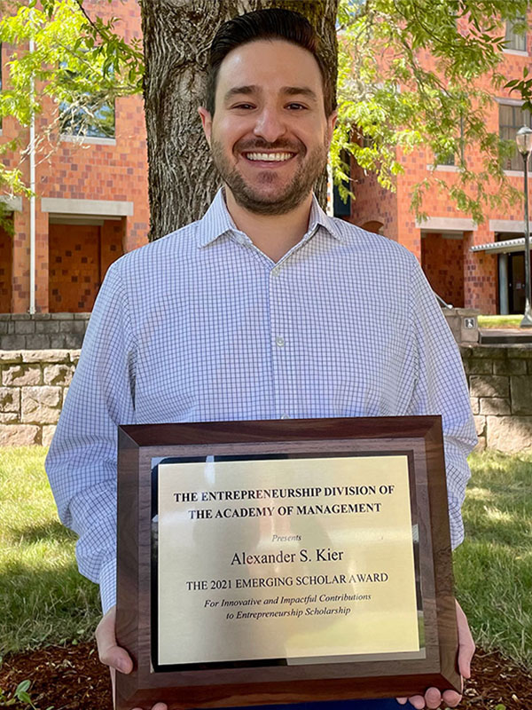 Alex Kier holding a framed award plaque from the Academy of Management’s Entrepreneurship Division, standing outdoors in front of a tree and brick campus buildings.