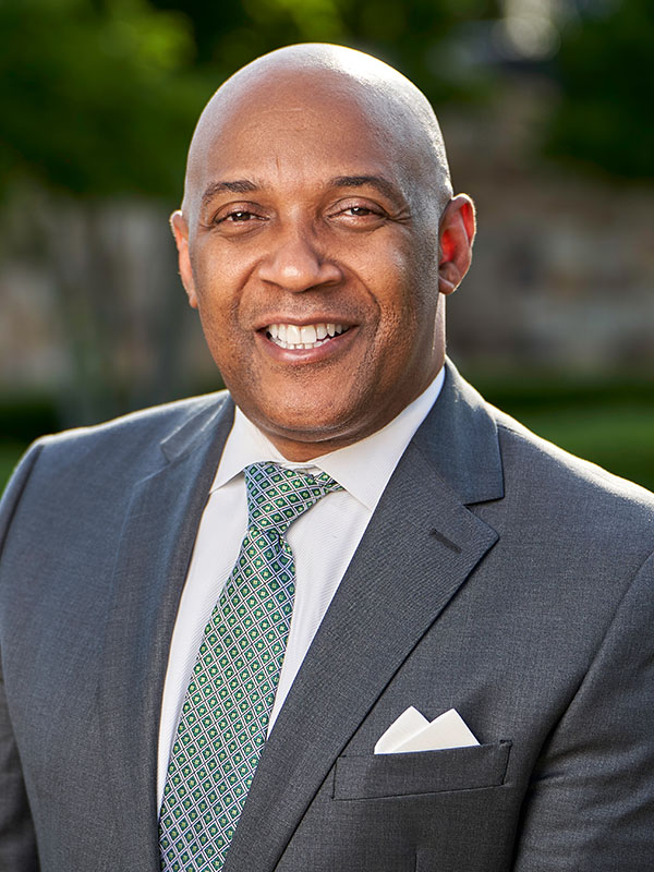 Bennie Harris wearing a gray suit with a patterned green tie and pocket square, standing outdoors with soft greenery in the background.