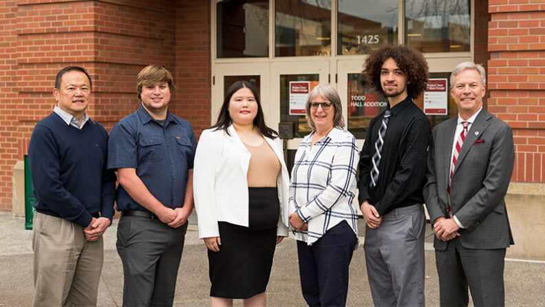 Zachary Sims, Mengdi Wei, and Ryan Smith standing with Accounting Chair Bernie Wong‑on‑Wing, Career Advisor Marla Meyer, and Dean Chip Hunter in front of a brick building entrance.