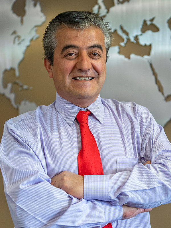 Dogan Gursoy standing with arms crossed, wearing a light purple dress shirt and bright red tie, posed in front of a world map wall display.