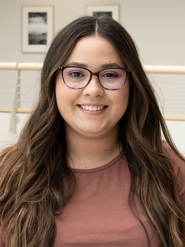 Flor Larios Ramos with long wavy brown hair, wearing a mauve top, standing indoors in a bright space with framed artwork and a railing in the background.