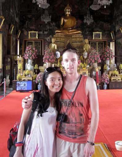 Jenny Trujillo and her husband, Tayvin, standing together inside an ornate Bangkok temple, with a large golden Buddha statue and floral arrangements behind them.