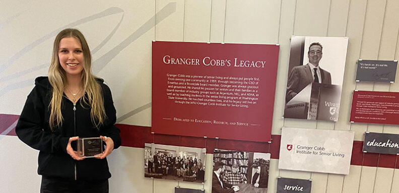 Kate Simonson stands in front of a Granger Cobb’s Legacy display wall, holding the Mather Institute Promising Practices Award, with photos and text panels behind her.