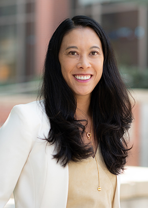 Hana Johnson wearing a cream blouse and white blazer, with long dark wavy hair, standing outdoors in front of softly blurred campus buildings.