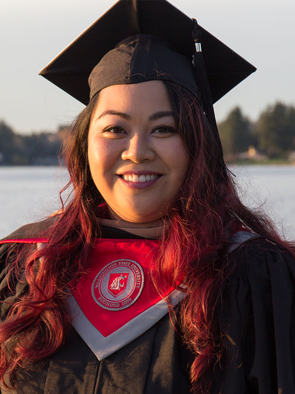 Hazelmae Overturf in a graduation cap and gown with crimson‑tinted hair, standing outdoors near a body of water at sunset, wearing a WSU graduation stole.