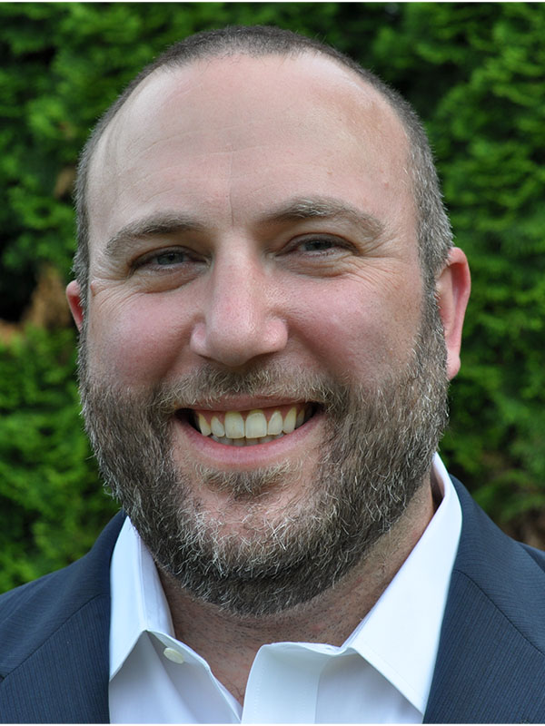 Jason Lee wearing a dark suit jacket and white collared shirt, standing outdoors in front of dense green foliage.