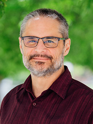 Jeff Joireman wearing a burgundy button‑up shirt, standing outdoors with bright green trees softly blurred in the background.