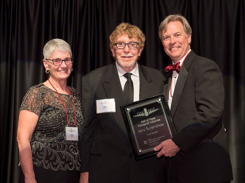 Nancy Swanger, Jerry Burtenshaw, and Dean Chip Hunter dressed in formal attire pose together onstage, with Burtenshaw holding a framed Hall of Fame award.