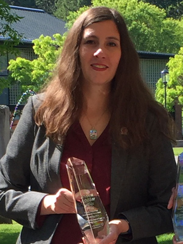 Jessica Aguilar holding a clear crystal award while standing outdoors in a sunlit garden area with trees and buildings in the background.
