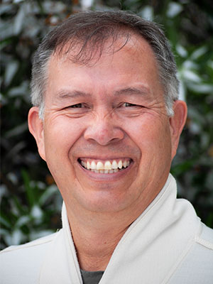 Joseph Scott Gladstone wearing a light-colored zip‑up jacket, standing outdoors in front of leafy green foliage.