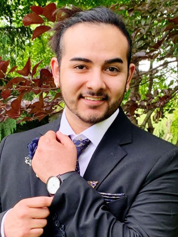 Larenze Rios adjusting his patterned tie while wearing a black suit and watch, standing outdoors beneath leafy trees.