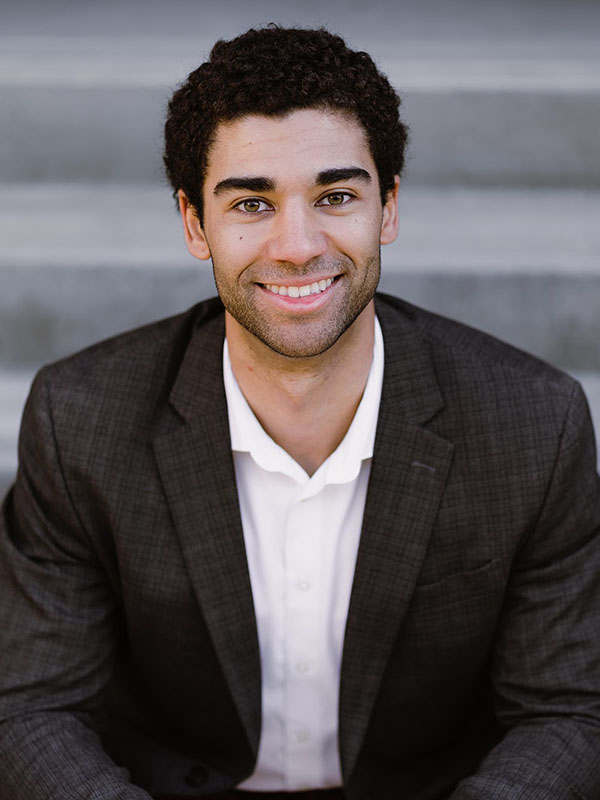 Marcus Lathan wearing a dark plaid suit jacket and white shirt, seated on outdoor concrete steps.